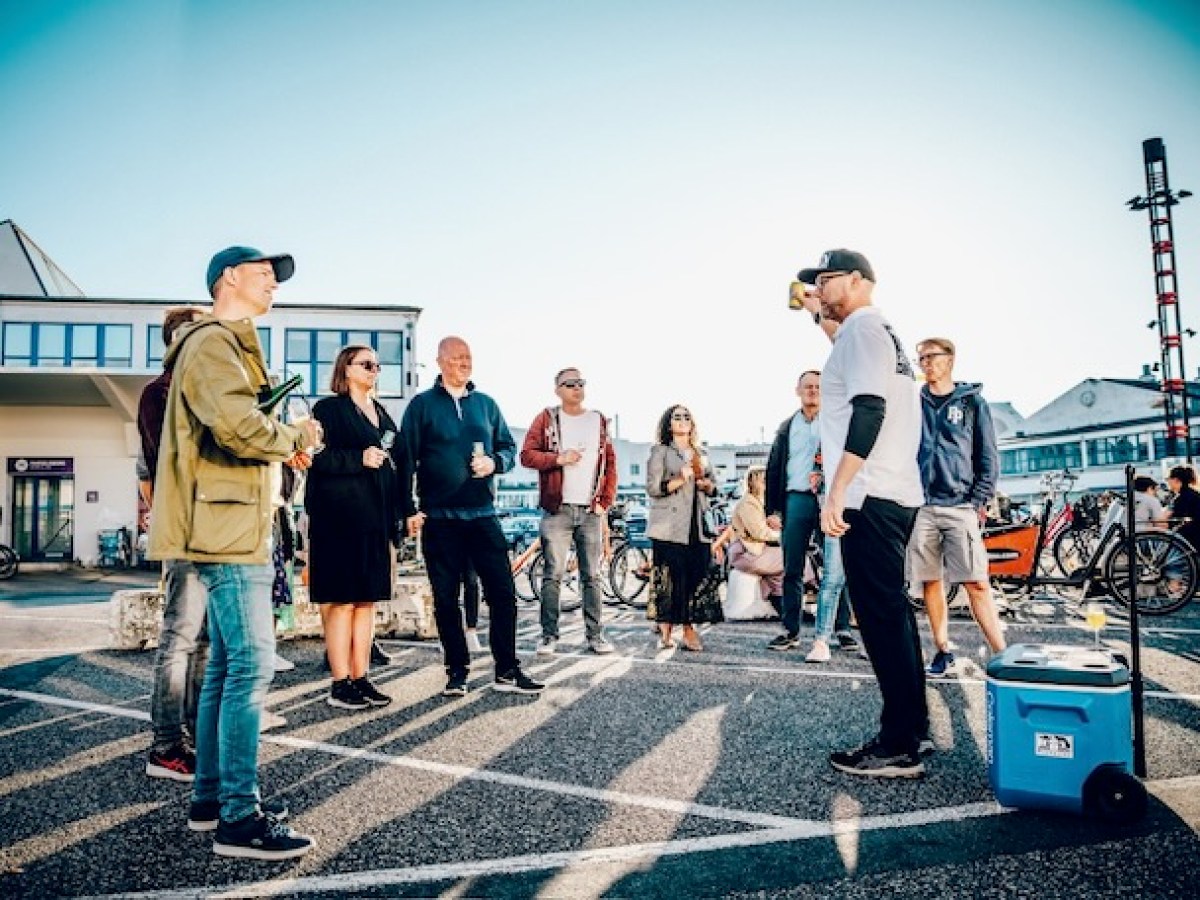 A group of tourists enjoying a BeerWalk in Copenhagen on a sunny summer day, listening to a local guide while tasting Danish craft beer outdoors.