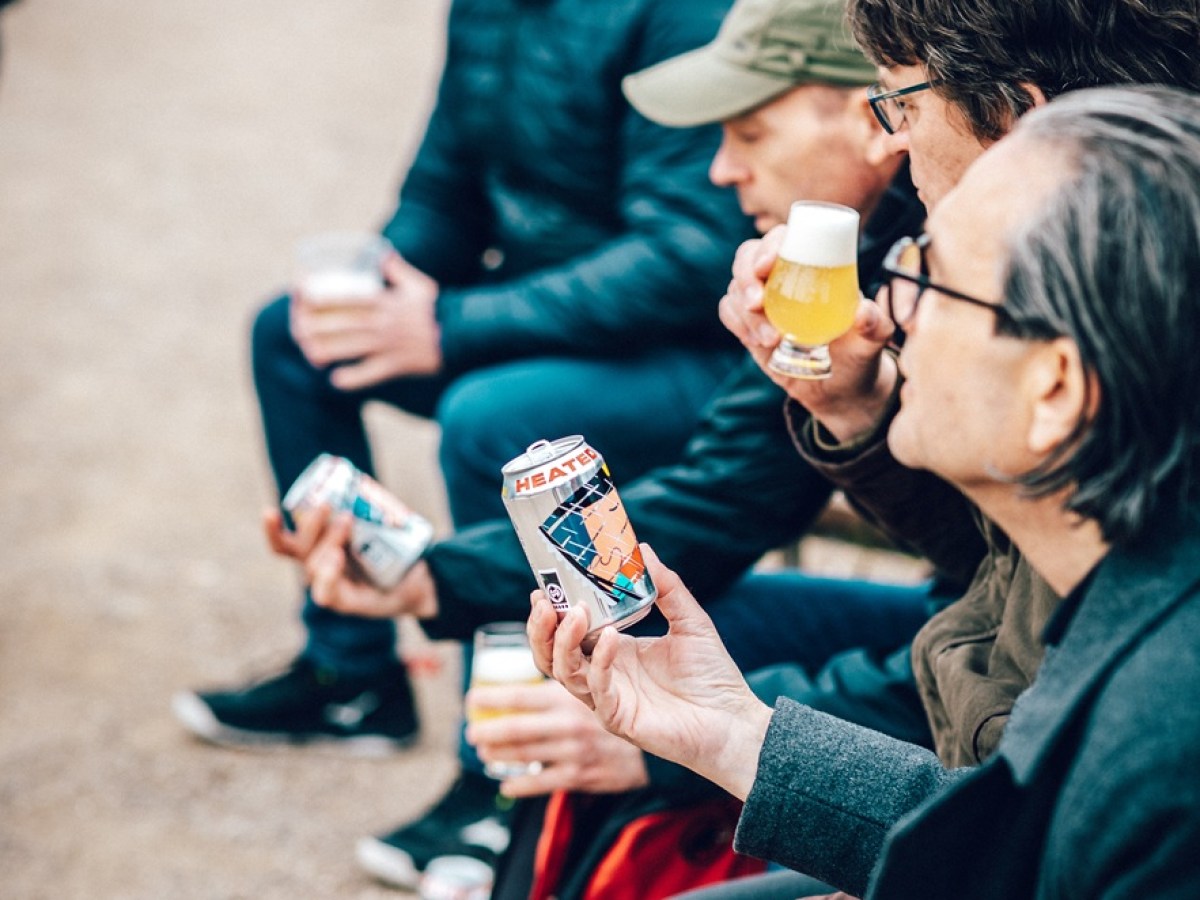 Four people sitting outdoors, holding and examining cans of beer.