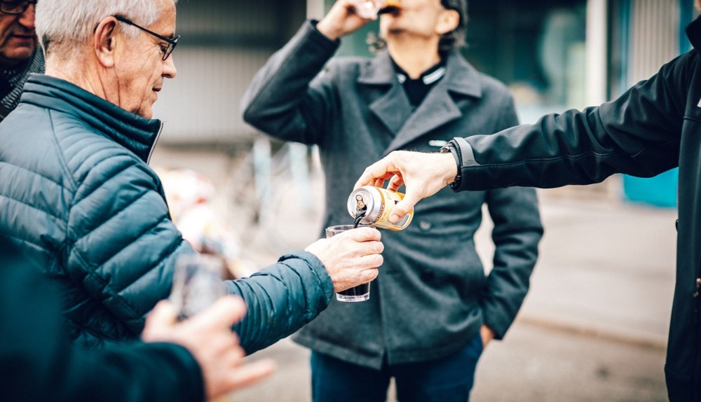 People outdoors sharing a drink, with a hand pouring from a can into a glass.