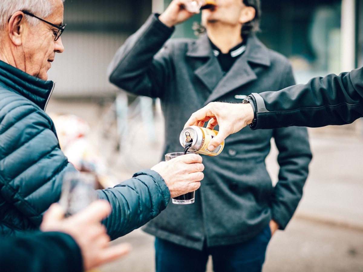 People outdoors sharing a drink, with a hand pouring from a can into a glass.