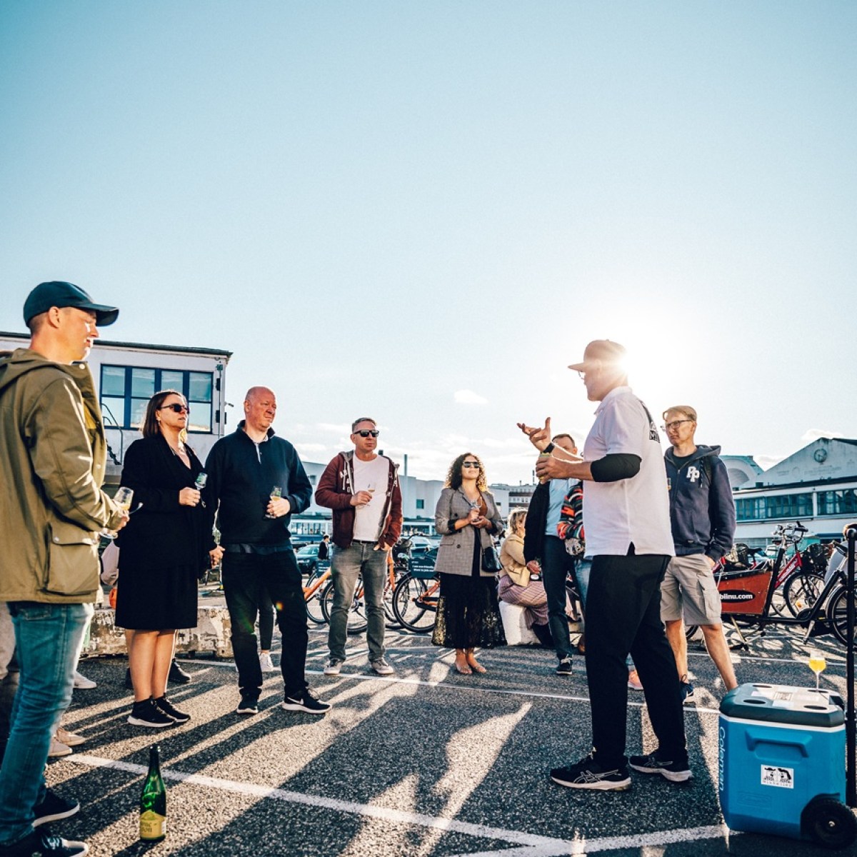 Group of people standing outdoors, listening to a speaker, with a blue sky background.