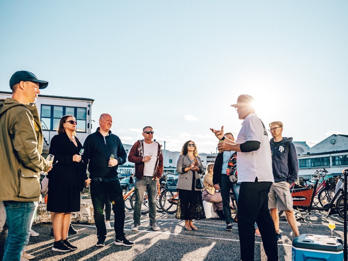 Group of people standing outdoors, listening to a speaker, with a blue sky background.
