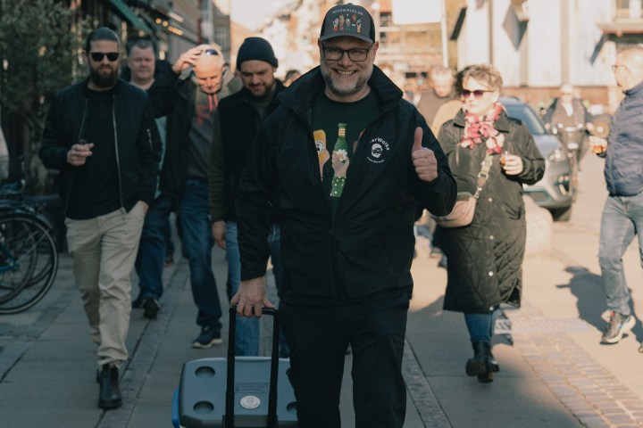 Man in cap pulling blue cooler, walking on sunny street with people in background.