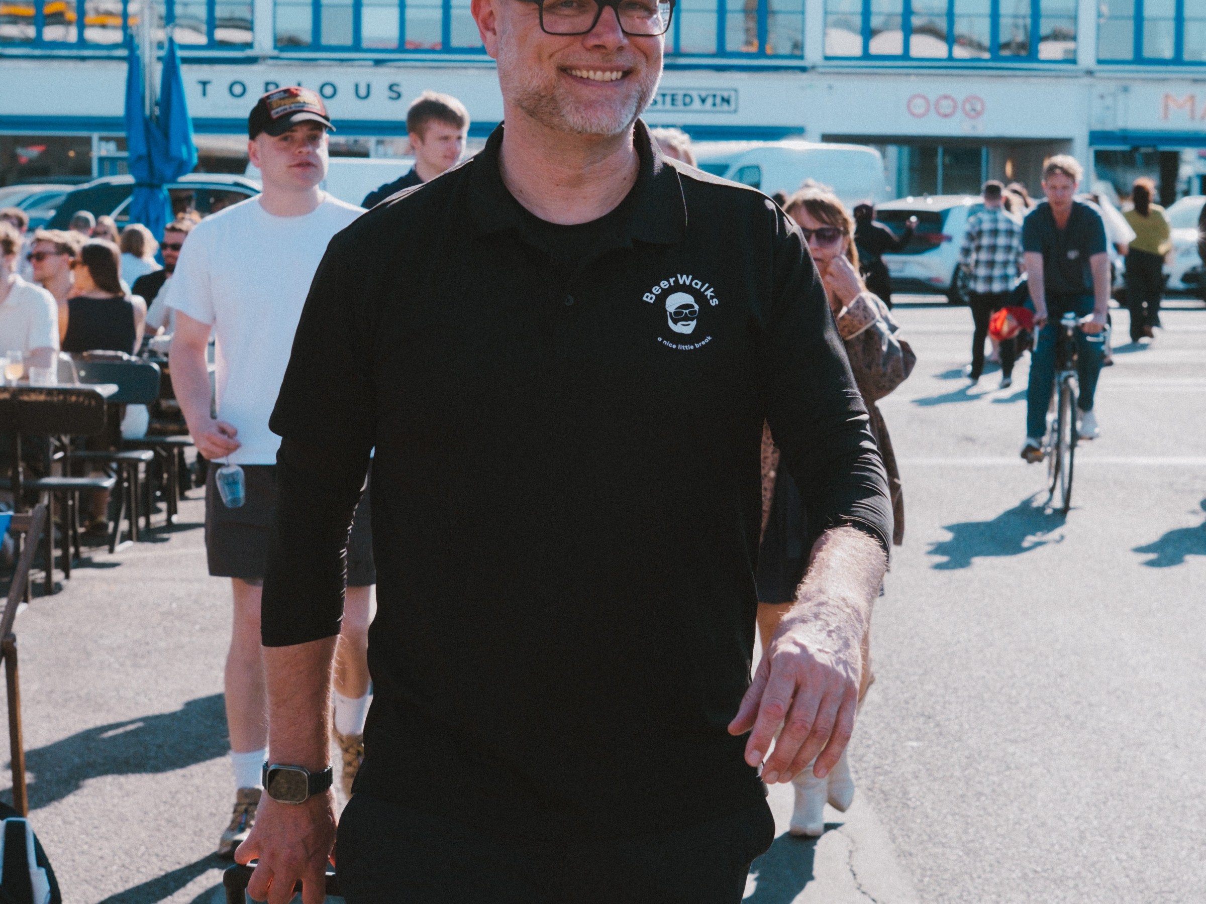 Man in black outfit smiling, pulling a cooler in a busy outdoor area.