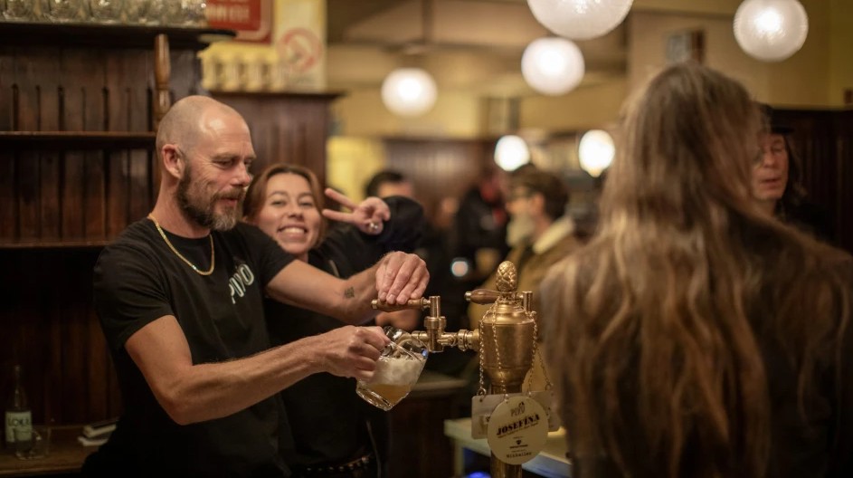 Bartender pouring beer with smiling woman posing behind him in a busy pub.